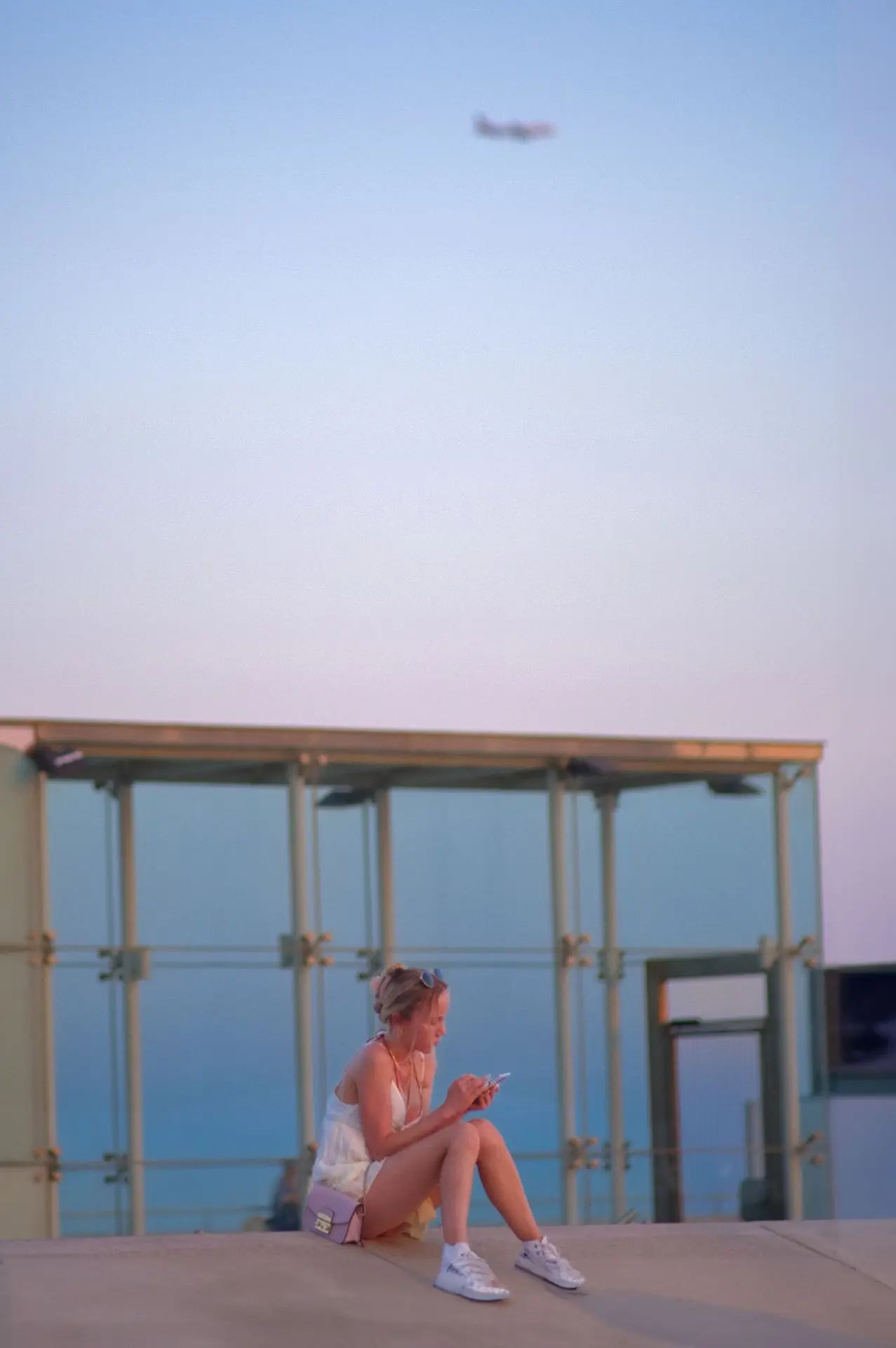 Seated before an endless view, a woman seems lost in her screen, while the glass architecture reflects the gentle hues of twilight. A scene capturing urban solitude and the beauty of fleeting moments in a city where every detail becomes a work of art.