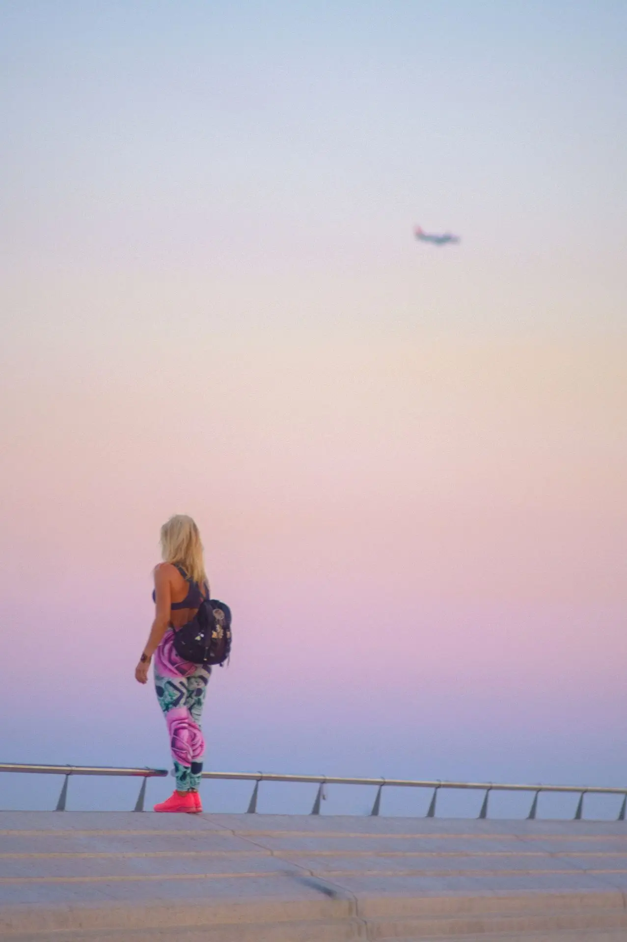 A woman observes the horizon from an elevated viewpoint, under a sky fading into soft pastel hues. The airplane in the background evokes a sense of travel and freedom, while the composition plays with perspective and contemplative solitude.