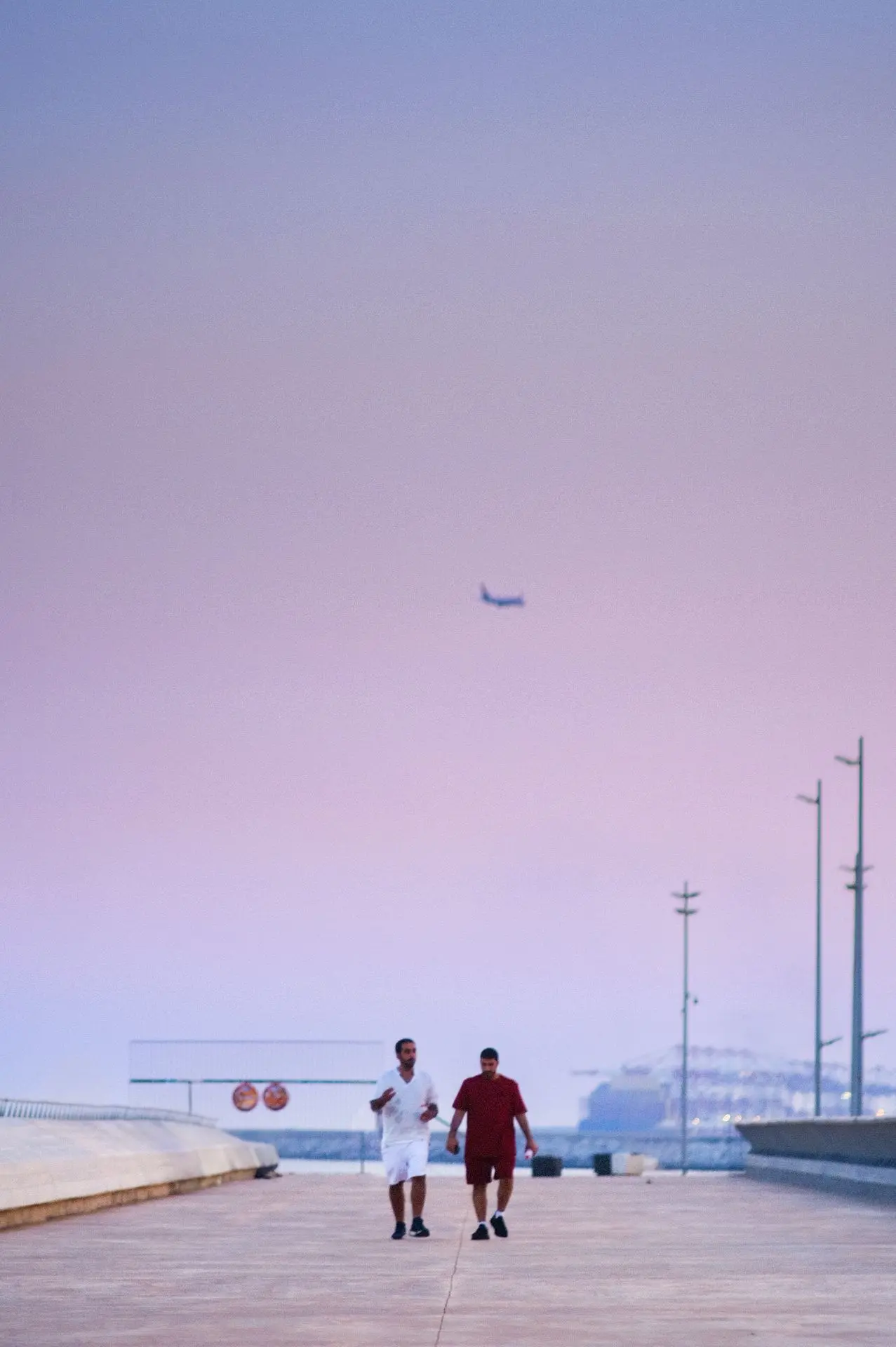 Two men walking through a minimalist setting, where the pastel sky blends seamlessly with the maritime horizon. A scene that captures the tranquility and energy of a late afternoon in Barcelona, balancing modern architecture with human connection.