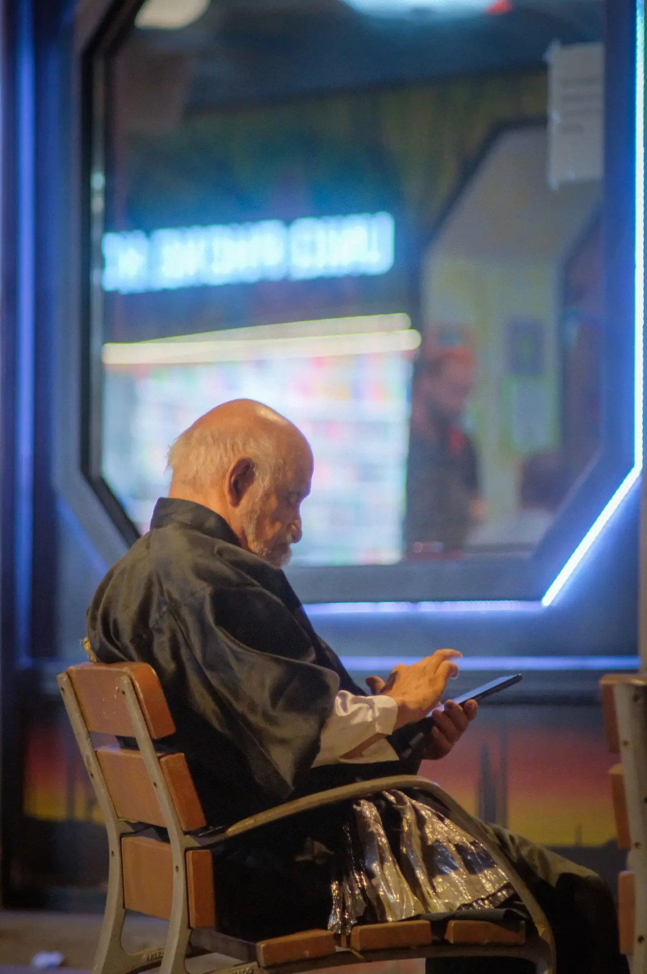 This image captures a modern urban moment, where technology and atmosphere merge in a striking visual composition. The subject, immersed in his phone, reflects the contemporary rhythm of life, where digital interactions often take precedence over the surrounding world. The neon lights behind him create a cinematic mood, bathing the scene in vibrant hues that contrast with his focused demeanor. The interplay of artificial light and shadow adds depth, evoking a sense of introspection and solitude within the bustling cityscape of Barcelona.