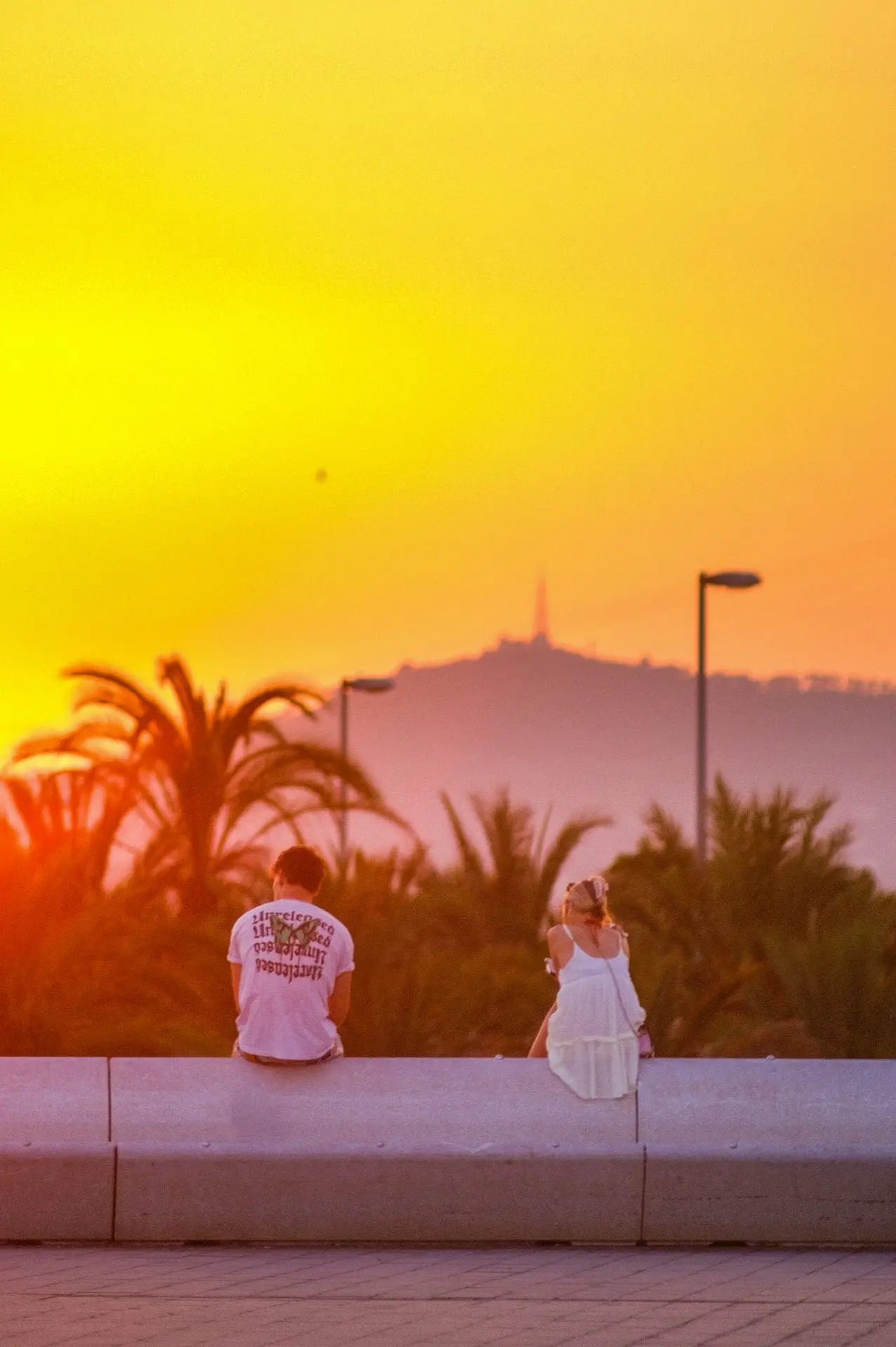 A scene capturing the magic of a Barcelona sunset, where two silhouettes gaze at the fiery horizon. The warm atmosphere and depth of field create a poetic visual composition, embodying the essence of travel and urban contemplation.